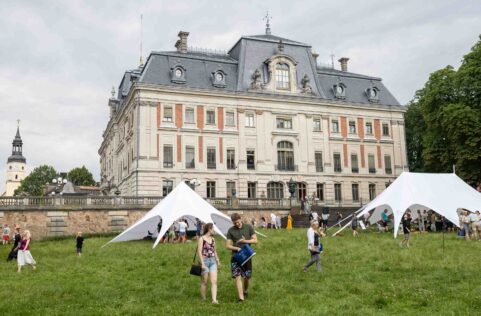 Szeroki kadr plenerowy przedstawiający parę młodych ludzi spacerujących po zielonej trawie na tle monumentalnego, zabytkowego Pałacu w Pszczynie. Na trawniku rozstawione są duże, białe namioty imprezowe w kształcie gwiazd, pod którymi gromadzą się uczestnicy rodzinnego pikniku.