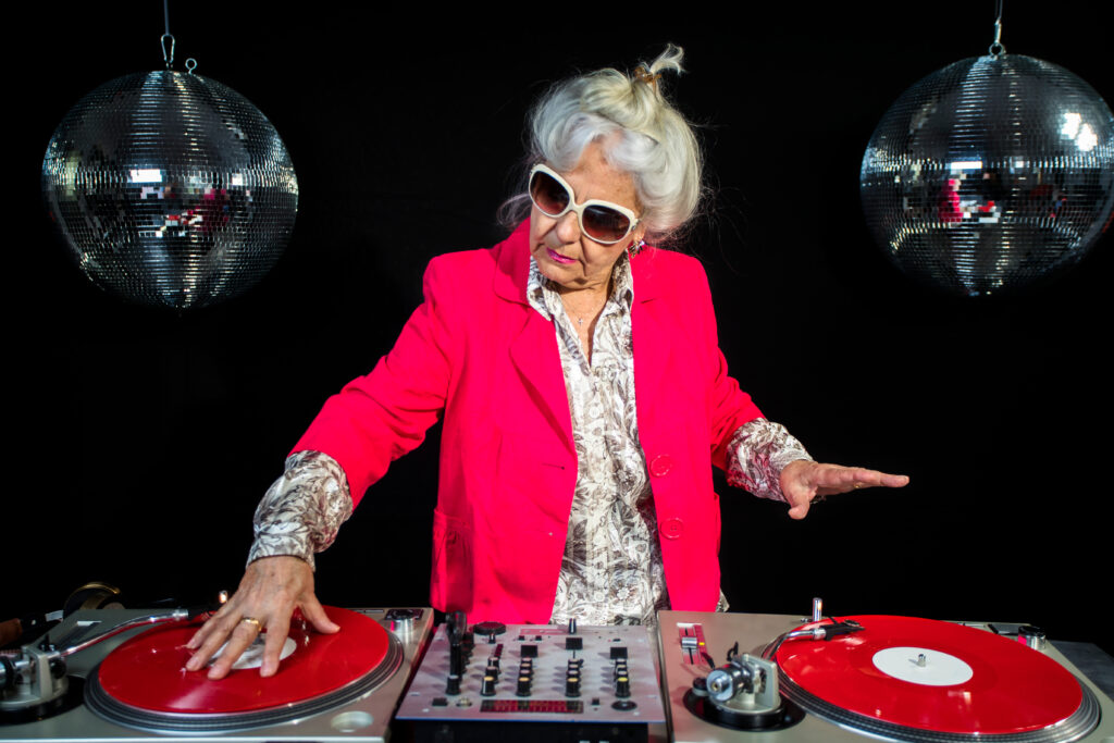 Starsza kobieta z siwymi włosami, ubrana w różową marynarkę i stylowe okulary przeciwsłoneczne, miksuje muzykę jako DJ. Trzyma dłoń na czerwonej płycie winylowej umieszczonej na gramofonie, stojąc skupiona przed profesjonalną konsolą mikserską. Ciemne tło zdobią dwie wiszące po bokach, błyszczące kule dyskotekowe, które podkreślają klubowy, pełen energii charakter sceny.