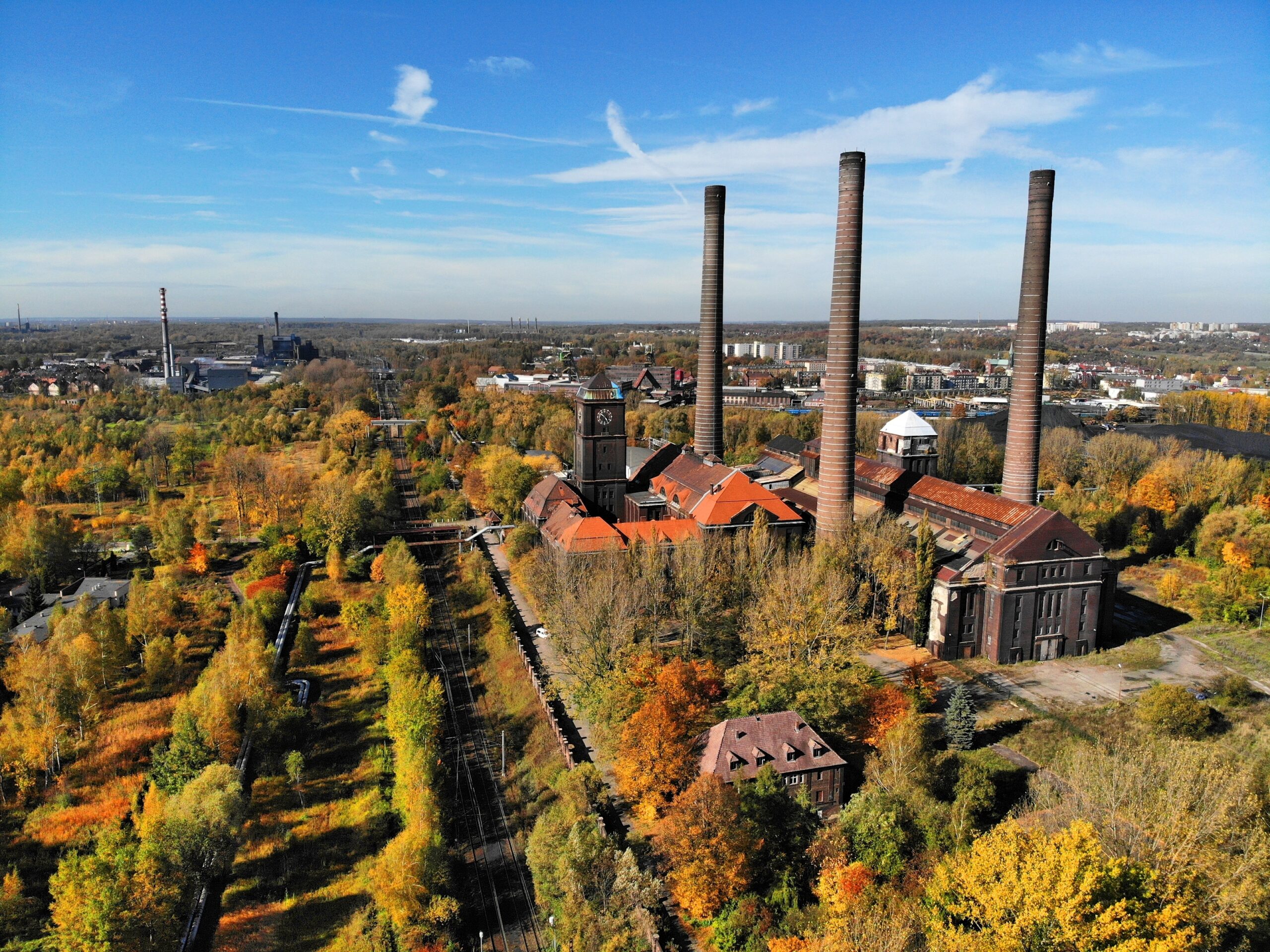 Widok z lotu ptaka na Bytom. Na pierwszym planie widoczny jest zabytkowy kompleks przemysłowy Elektrociepłowni Szombierki z ceglanymi budynkami o pomarańczowych dachach i trzema monumentalnymi, wysokimi kominami. Obiekt otoczony jest drzewami w jesiennych barwach, a tuż obok niego przebiegają tory kolejowe. W oddali, pod bezchmurnym niebem, rozciąga się panorama miasta wraz z terenami poprzemysłowymi i przemysłowymi.