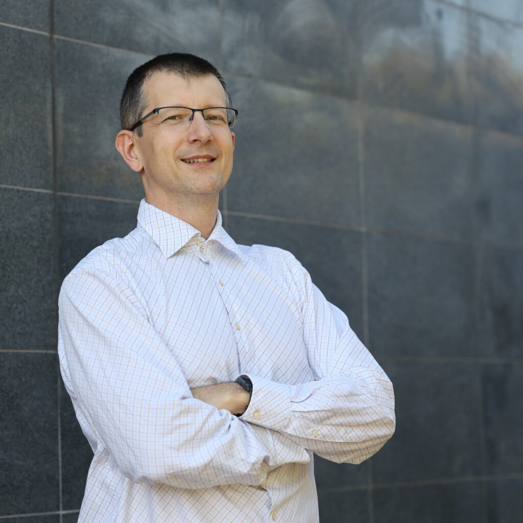 Ujęcie mężczyzny w okularach i jasnej koszuli w kratkę, stojącego ze skrzyżowanymi ramionami na tle nowoczesnej ściany z ciemnoszarych paneli. Mężczyzna ma krótkie, ciemne włosy i uśmiecha się lekko, patrząc w kierunku obiektywu; na jego lewym nadgarstku widoczny jest zegarek. Oświetlenie jest naturalne i równomierne.