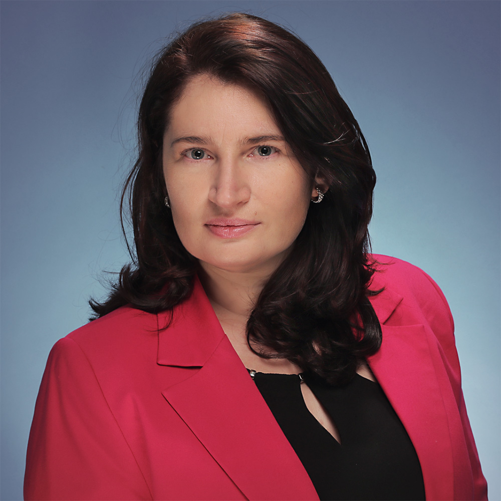 Biznesowy portret kobiety o ciemnobrązowych, pofalowanych włosach sięgających ramion, która patrzy spokojnie w stronę aparatu. Ma na sobie wyrazisty żakiet w kolorze fuksji założony na czarną bluzkę oraz subtelne kolczyki. Tło zdjęcia jest w pełni jednolite, utrzymane w stonowanym, niebiesko-szarym gradiencie.
