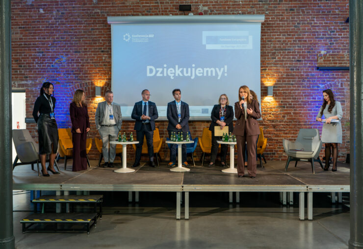 Zdjęcie przedstawia podsumowanie jednego z paneli dyskusyjnych na scenie w industrialnej hali o ceglanych ścianach. Grupa prelegentów stoi w linii na tle ekranu z napisem „Dziękujemy!”, a dyrektor Śląskiego Centrum Przedsiębiorczości Elżbieta Kabelis zabiera głos, przemawiając do mikrofonu. Obok nich znajdują się fotele oraz stoliki z napojami, wpisujące się w nowoczesno-przemysłową przestrzeń konferencyjną.